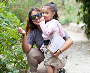A parent kneels and smiles near her child in a garden. The kid is eating something small and red, possibly a berry.