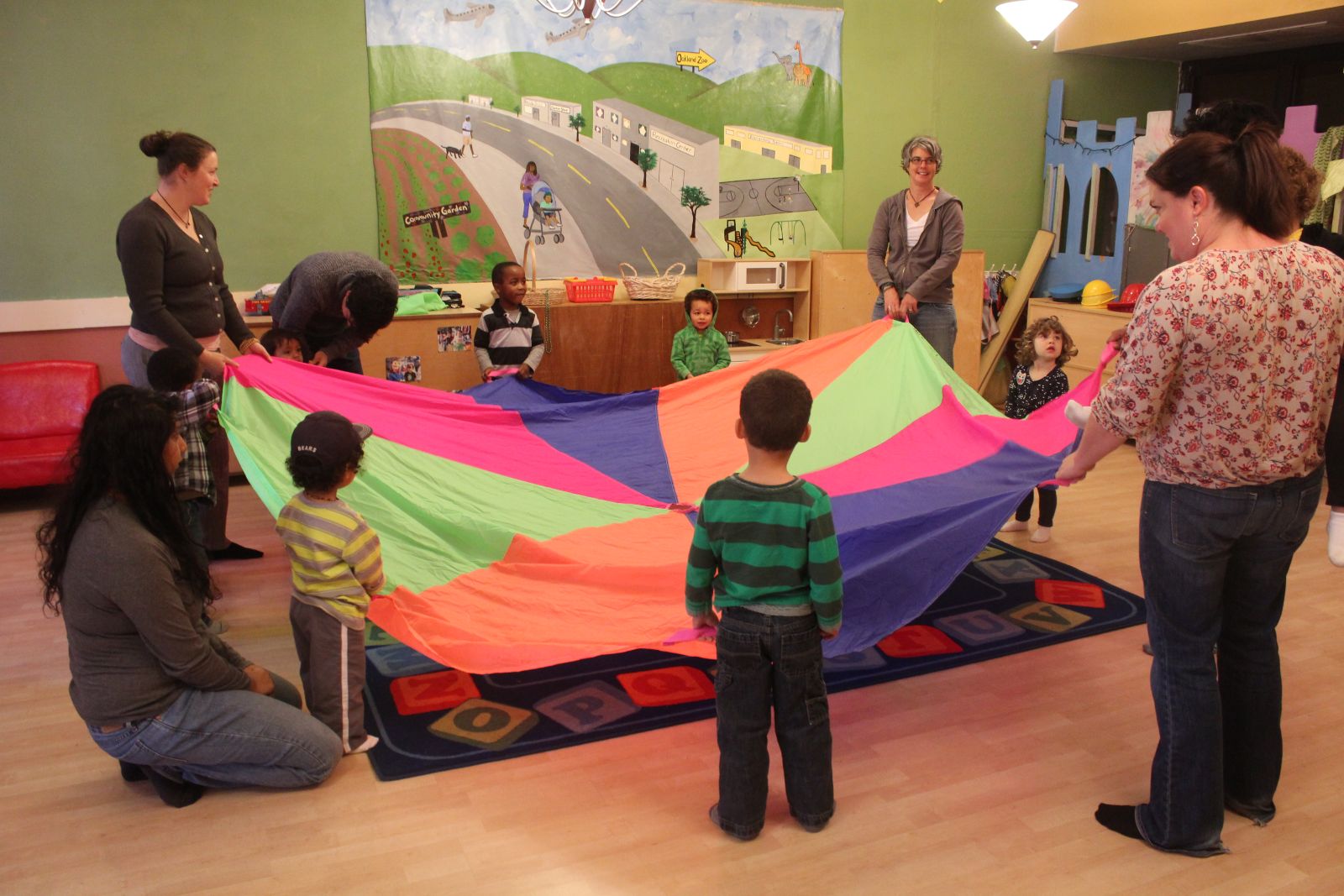 A diverse group of young children and their care givers hold up a big, colorful parachute. They are in a play space complete with a castle, a painting of a parent walking down the street with a baby in a stroller, and an alphabet rug.