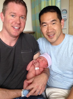 Two dads hold a newborn baby, still wearing a medical bracelet.