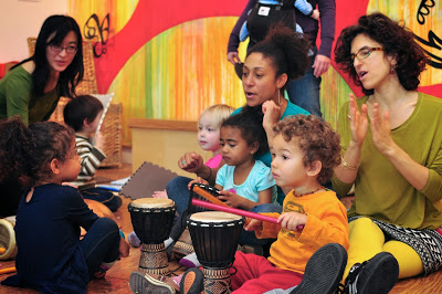 A diverse group of children and adults clap, sing, and play percussion instruments.