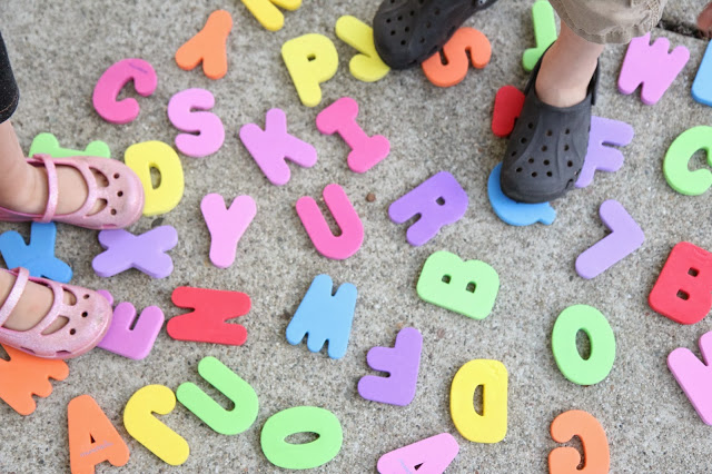 Two pairs of toddlers' feet, standing atop colorful foam letters, on asphalt. 