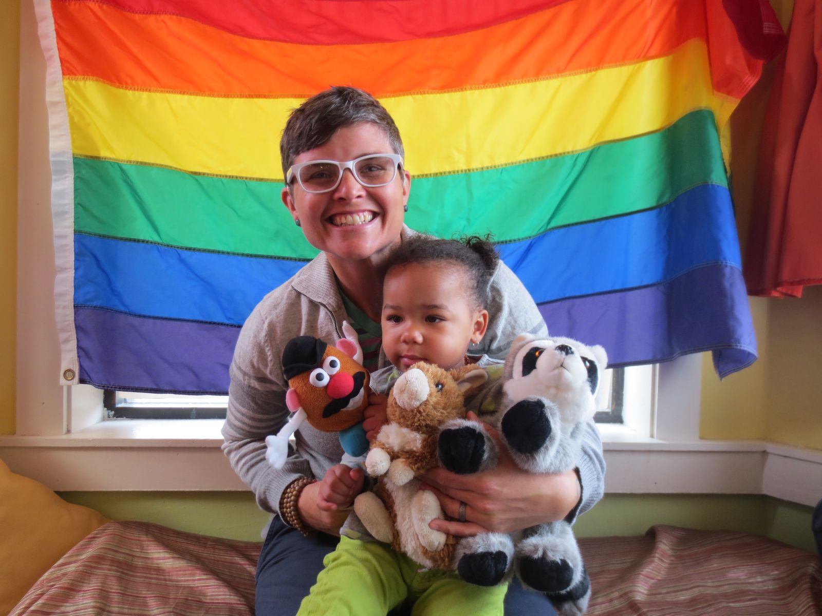 A smiling parent poses in front of a rainbow flag with their child, who is holding several stuffed animals.