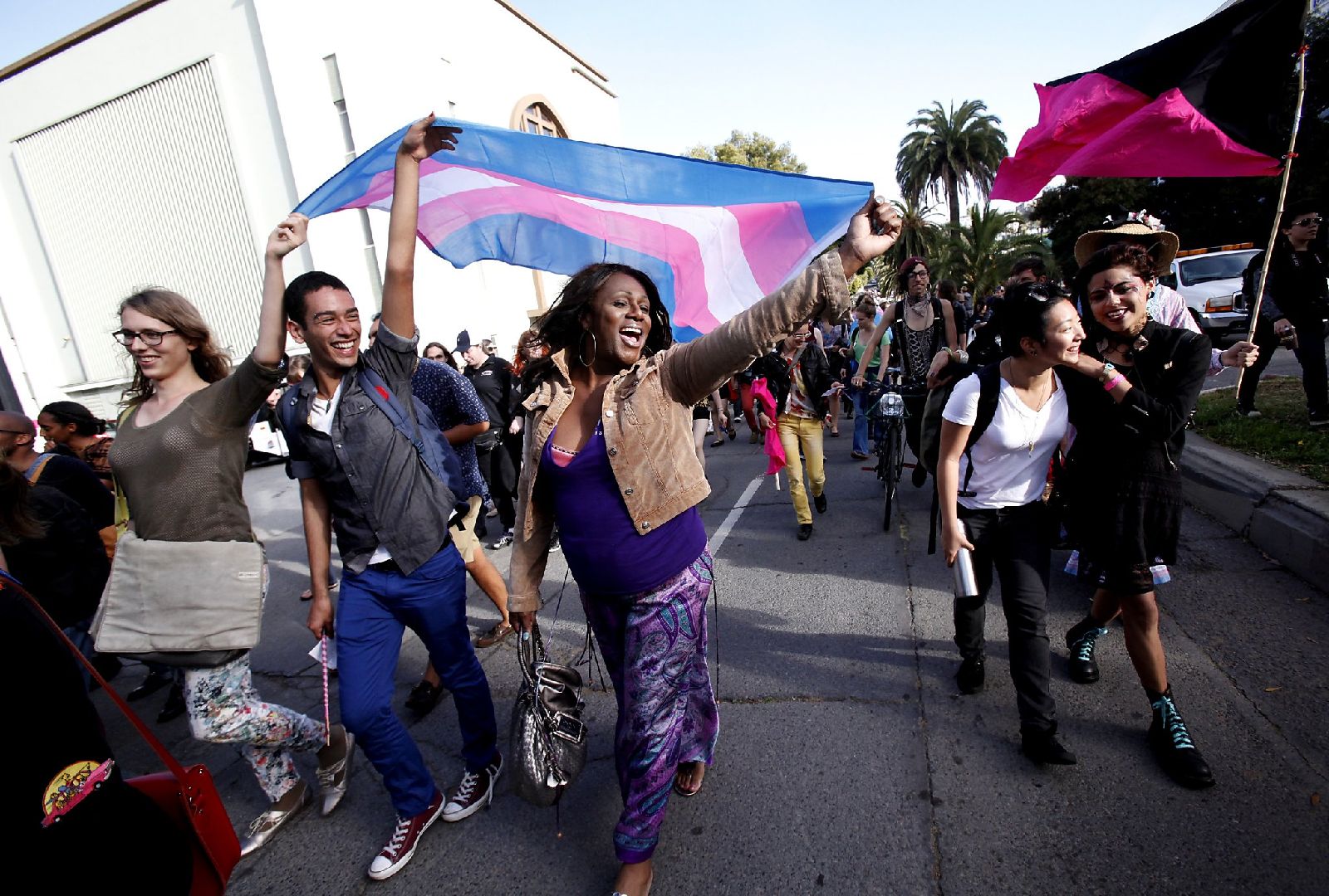 A crowd of folks march. Three people smile while holding up a trans flag that waves in the wind behind them.